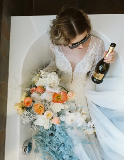 Coole Braut im Brautkleid mit Prosecco und Brautstrauß in einer Badewanne in ihrer Hochzeitslocation in den Bergen