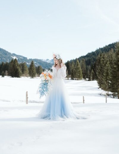 Braut beim Shooting auf ihrer Winterhochzeit in den Bergen mit Flowercrown und Brautstrauß umgeben von Bergpanorama