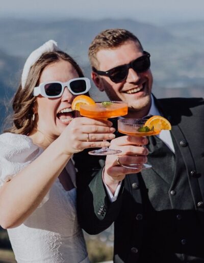 Glückliches Brautpaar auf ihrer Berghochzeit beim Anstoßen mit Aperol Spritz