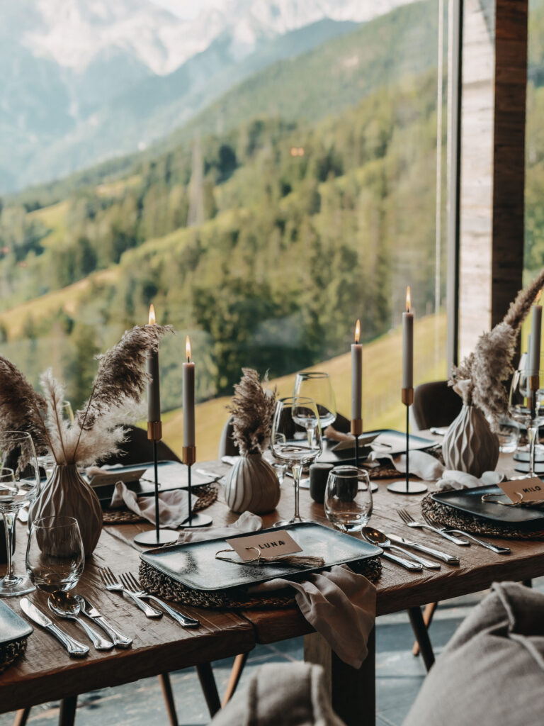 Elegante Tischdekoration mit Kerzen, Glaswaren und Pampasgras, mit Blick auf die üppig grüne Berglandschaft des Hochkönigs.