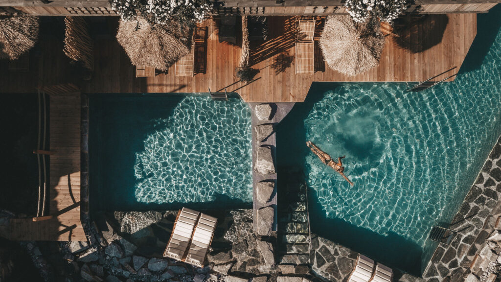 Eine Person schwimmt in einem klaren, blauen Pool, der von Holzterrassen und Liegestühlen umgeben ist – aus der Vogelperspektive betrachtet vor der atemberaubenden Kulisse des Hochkönigs.