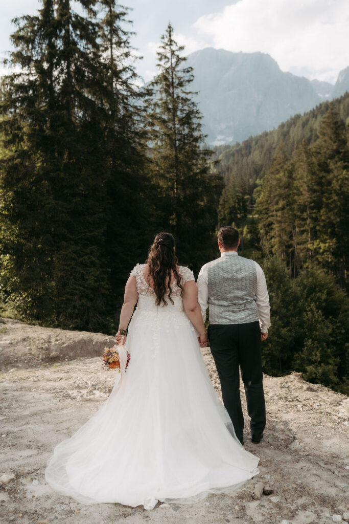 Braut und Bräutigam halten Händchen, blicken auf die atemberaubende Hochkönig-Berglandschaft und den Wald und halten ihren Hochzeitsstrauß in der Hand.