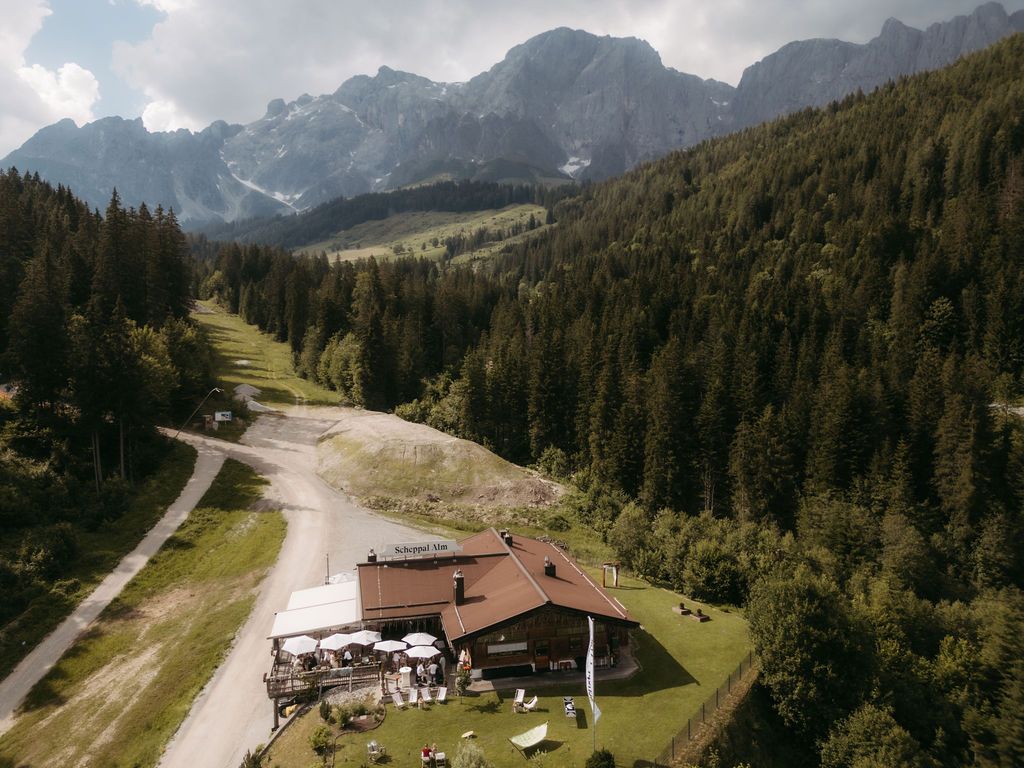 Berggasthof mit Sitzgelegenheiten im Freien, umgeben von Wald und Bergen unter teilweise bewölktem Himmel, mit atemberaubendem Blick auf den Hochkönig.