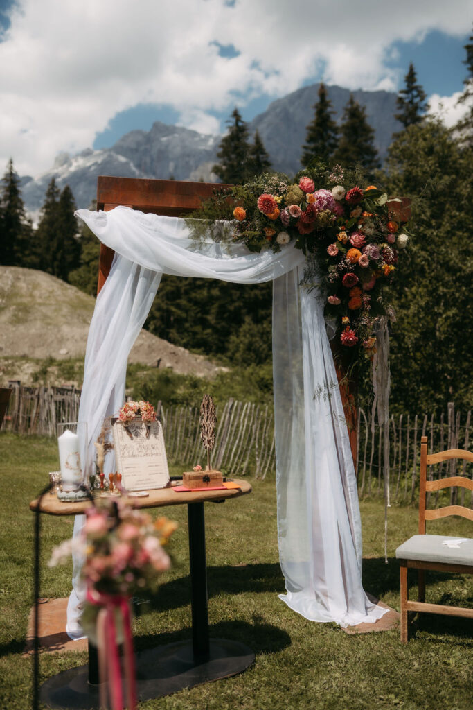 Im Freien steht ein Hochzeitsbogen mit Blumen und weißen Vorhängen, im Hintergrund erheben sich die majestätischen Hochkönig-Berge und der Wald.