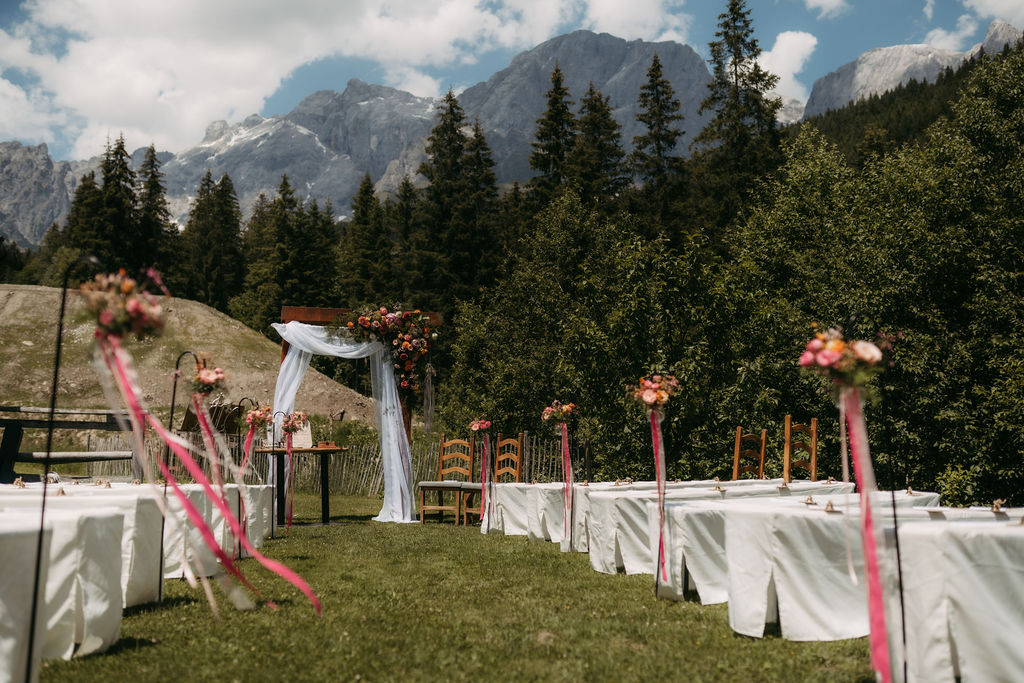 Hochzeitsdekoration im Freien mit weißen Stühlen, Blumenschmuck und Bergen im Hintergrund unter blauem Himmel, die einen atemberaubenden Blick auf den majestätischen Hochkönig bietet.