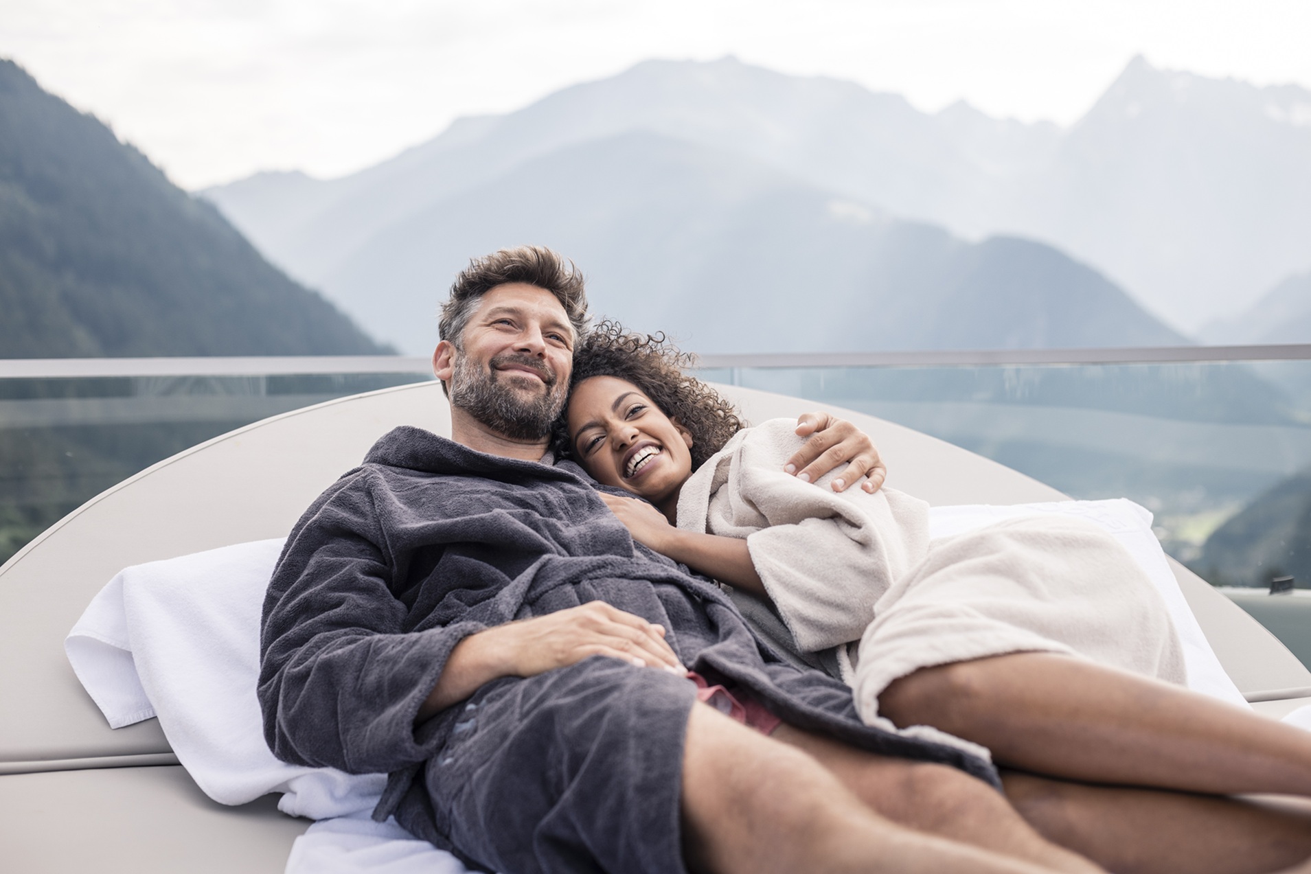 Verliebtes Paar in Bademänteln auf einer Relaxliege mit Ausblick auf die Alpen – Zweisamkeit und Erholung im Wellnesshotel für Paare in Tirol.