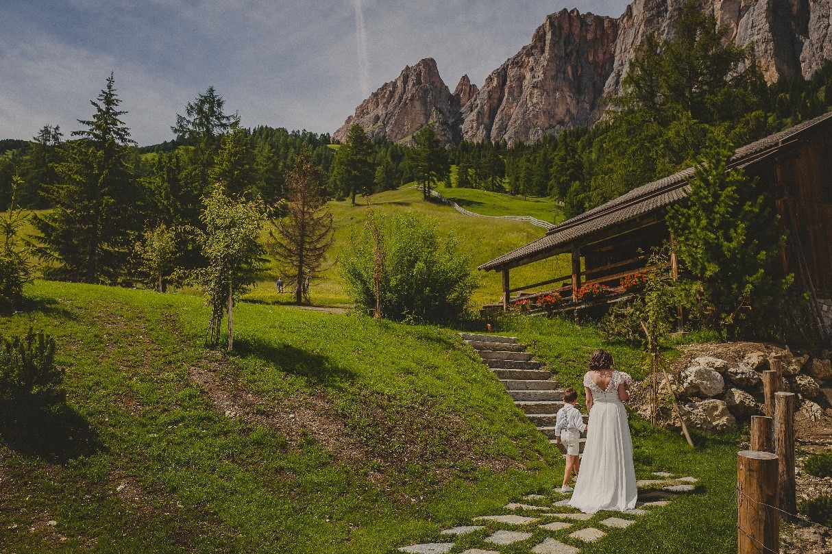 A woman in a white dress and a child walk up stone steps towards a cozy cabin surrounded by mountains - an idyllic mountain wedding venue.