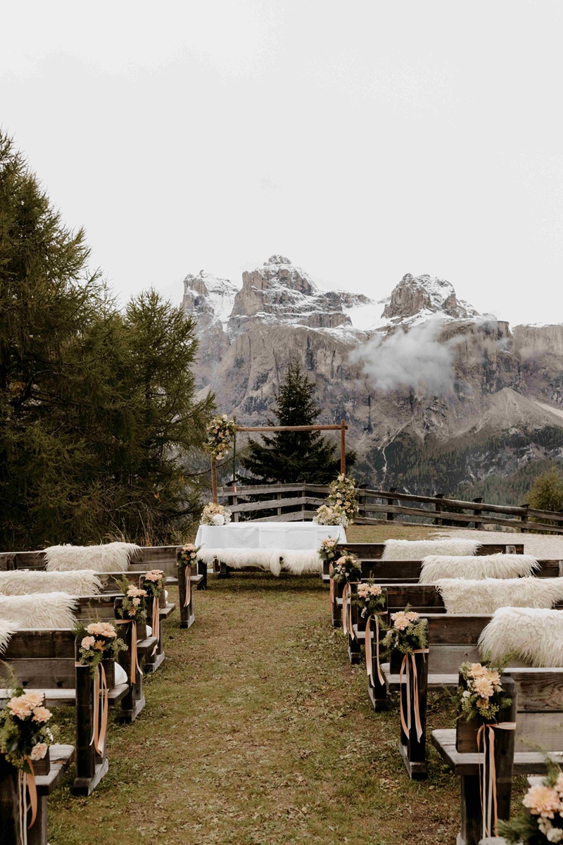 Trauung im Freien mit Blick auf die verschneiten Dolomiten – rustikale Bänke mit Fellen und Blumenarrangements für eine romantische Winterhochzeit in den Bergen.