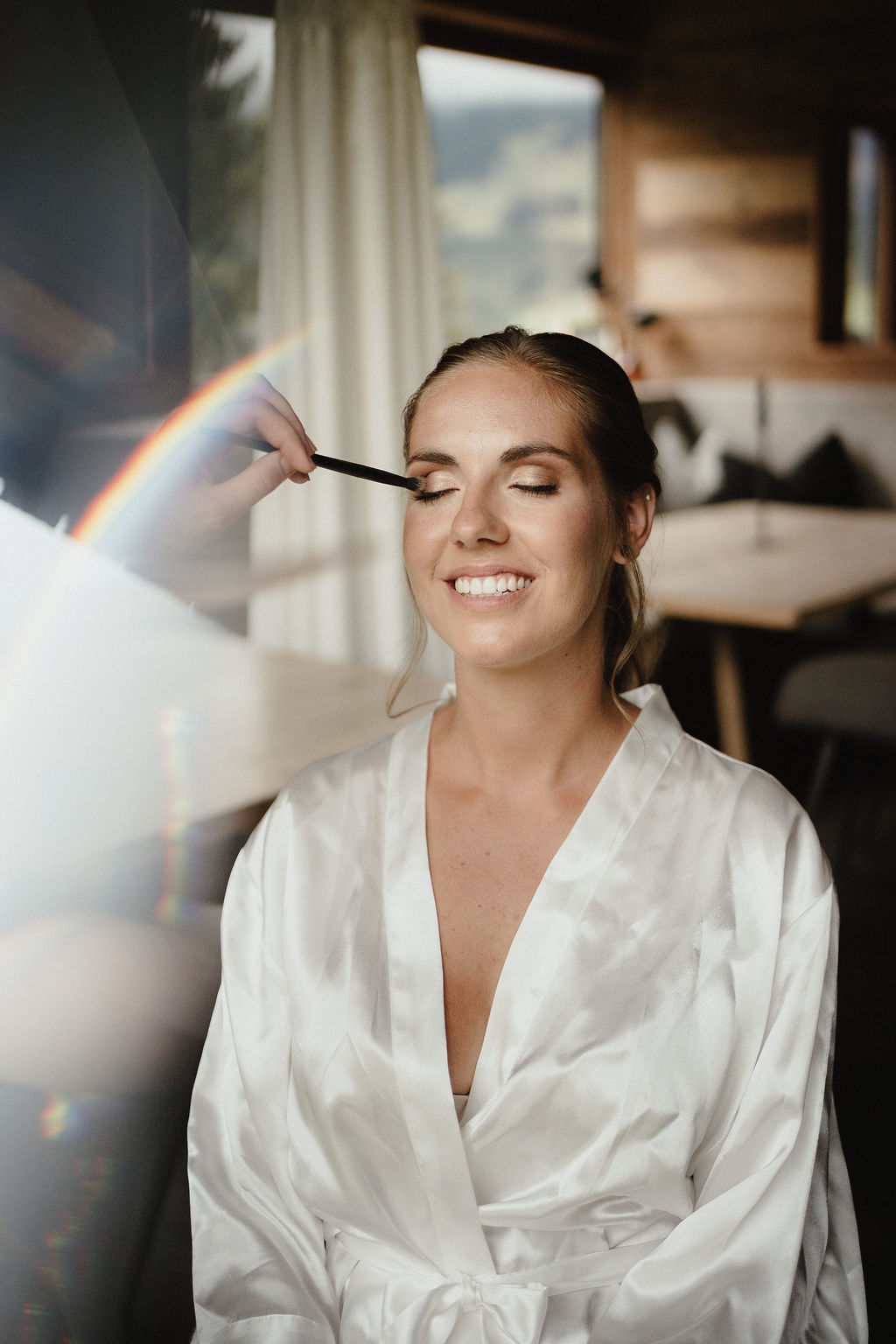 Eine lächelnde Frau in einer weißen Robe wird geschminkt, mit einem Regenbogenlicht im Vordergrund - perfekte Momente vor einer Berghochzeit in einer atemberaubenden Hochzeitslocation in Österreich.