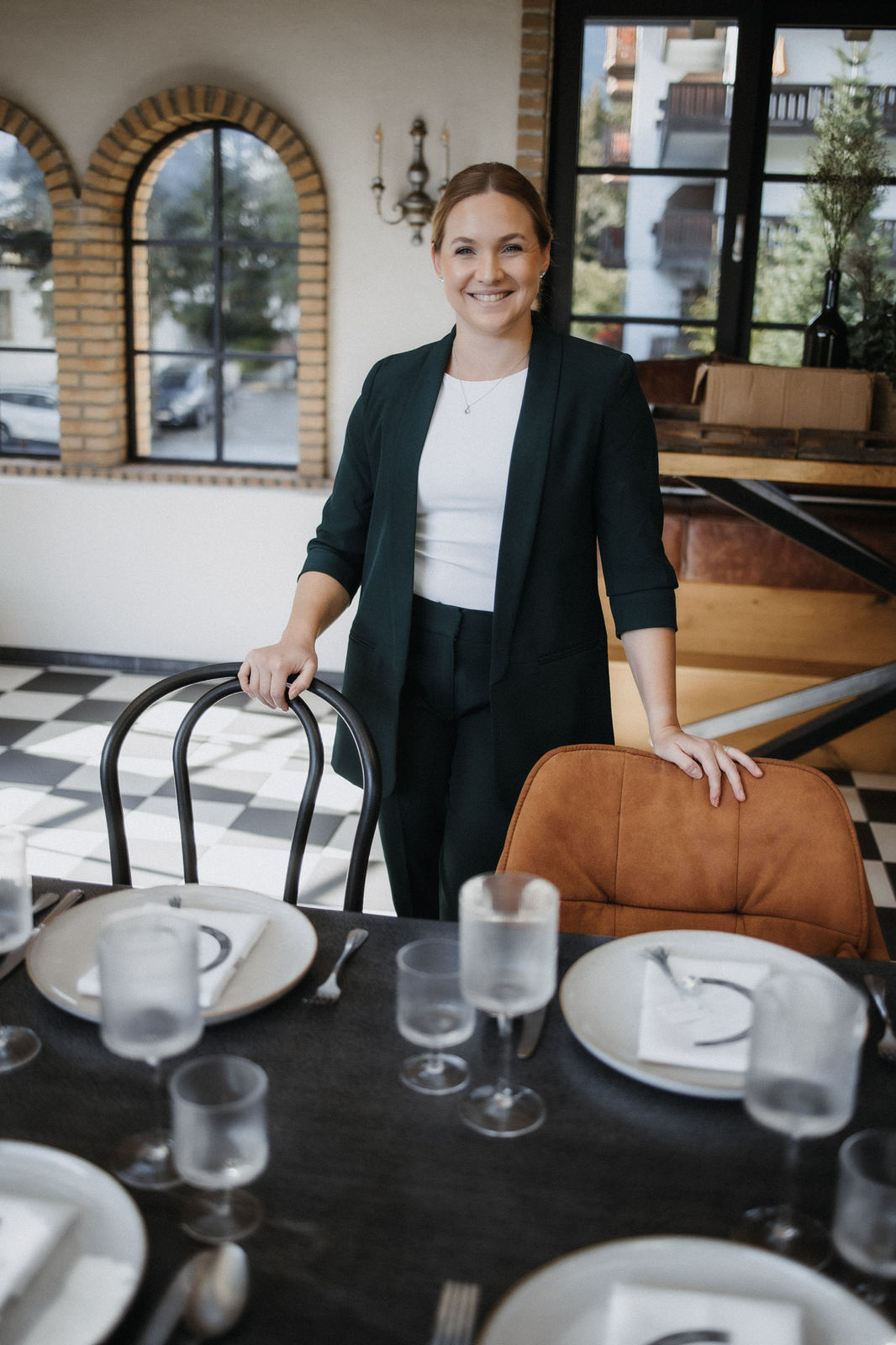 Ronja Baumbach in einem dunklen Anzug steht lächelnd an einem mit Tellern und Gläsern gedeckten Esstisch in einem hellen, stilvollen Raum - perfekt für eine intime Winterhochzeit oder eine elegante standesamtliche Trauung.