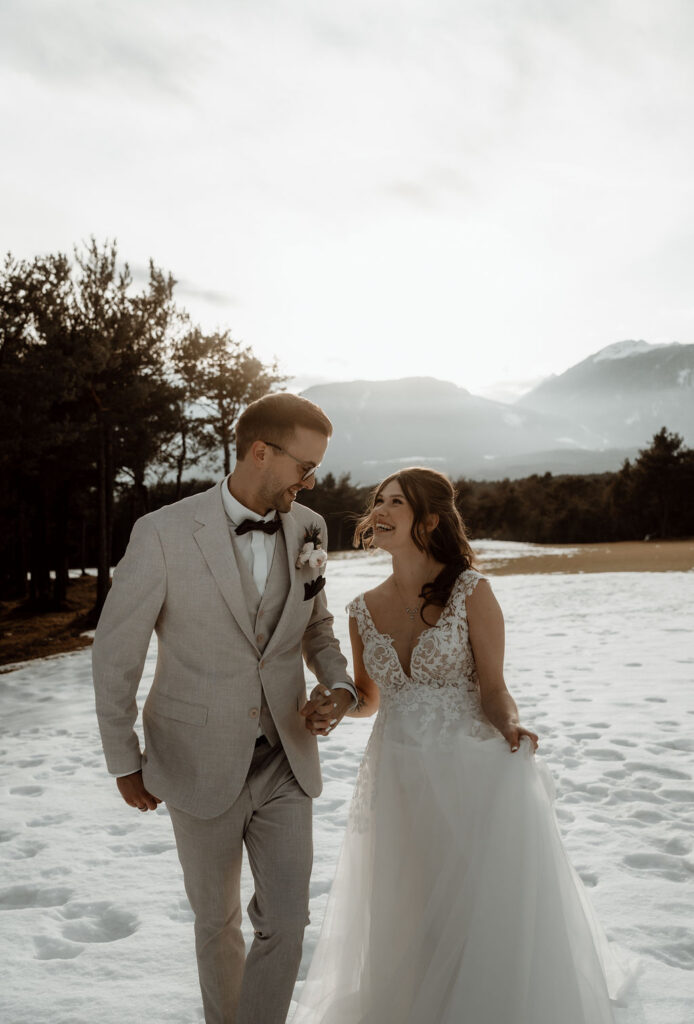 Braut und Bräutigam lächeln und halten sich an den Händen, während sie auf verschneitem Boden mit Bergen im Hintergrund spazieren gehen und den Zauber einer Winterhochzeit in einer atemberaubenden Hochzeitslocation festhalten.