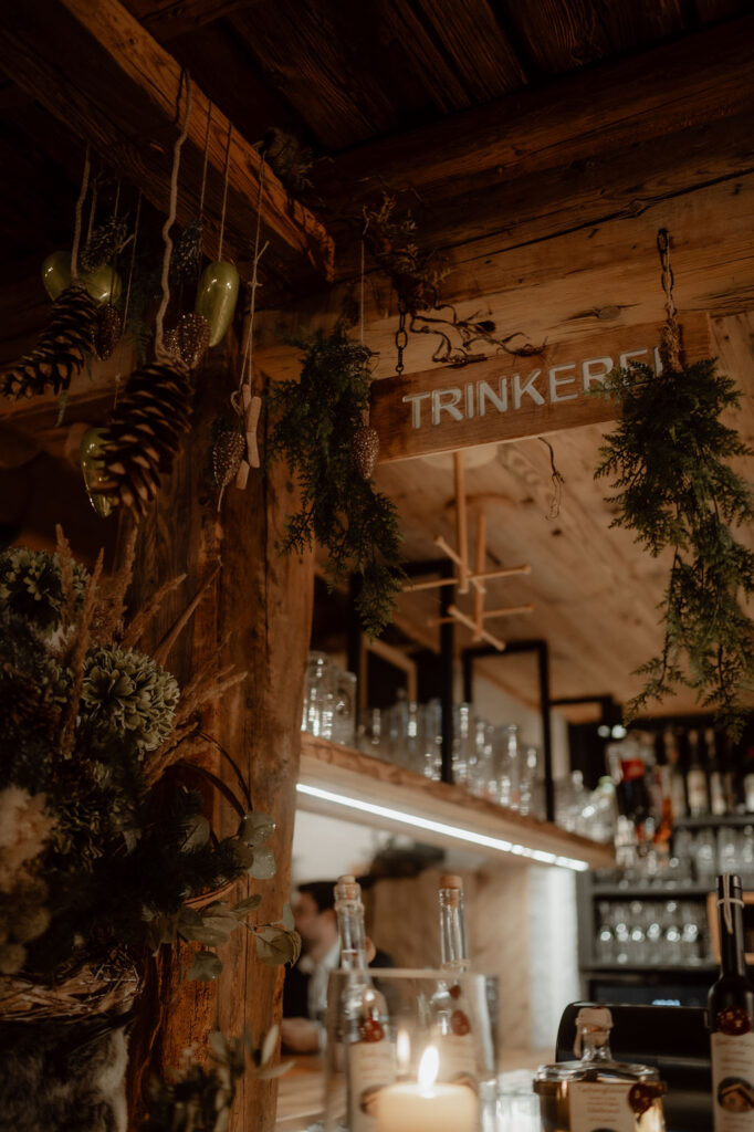 Rustic bar with wooden beams, greenery, pine cones, glasses, and a sign reading TRINKEREI - perfect for a mountain wedding or civil ceremony in Austria.
