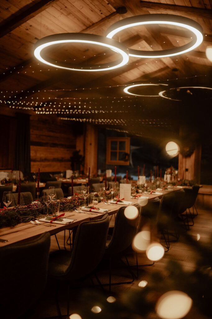 A warmly lit, rustic dining hall with long tables set for a festive winter wedding dinner, and modern circular lights above – a charming wedding venue for your mountain wedding celebration.