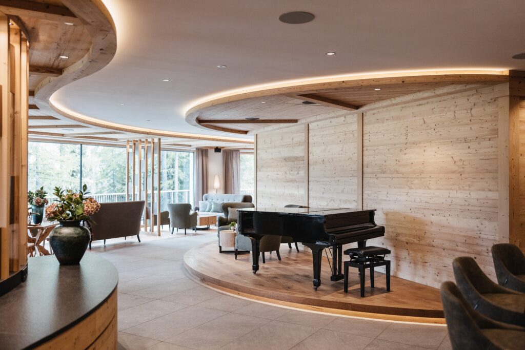 Moderne Lounge mit hellen Holzwänden, einem Flügel auf einer kleinen Bühne, Sofas und großen Fenstern mit Blick auf den Wald.