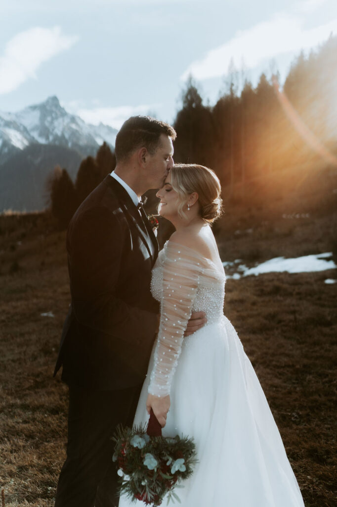 Der Bräutigam küsst die Stirn der Braut in einer atemberaubenden Tiroler Berglandschaft bei Sonnenuntergang; die Braut hält den Brautstrauß, beide lächeln - eine unvergessliche Berghochzeit in Österreich.