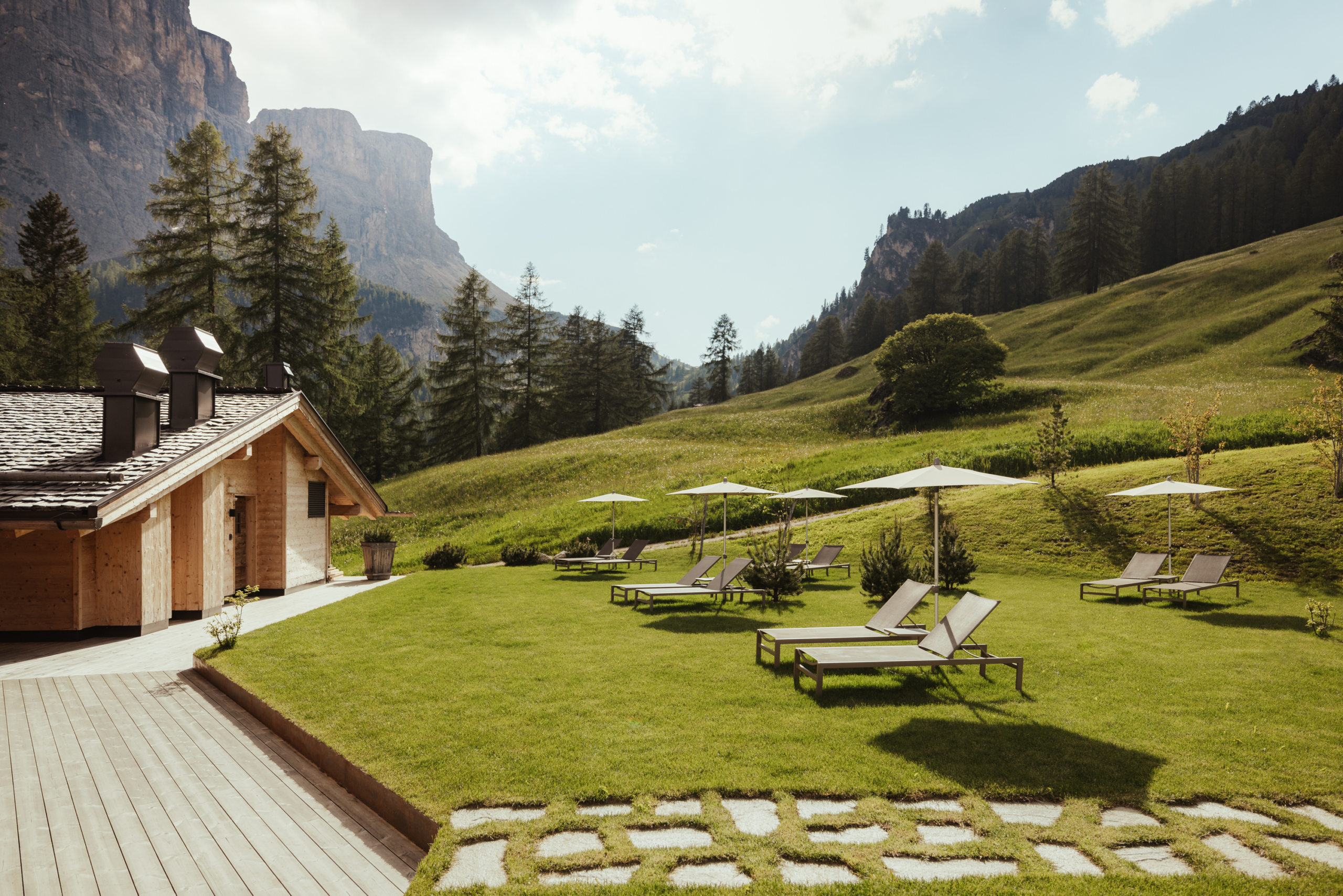 Lounge chairs and umbrellas on a meadow next to a wooden cabin with mountains in the background.