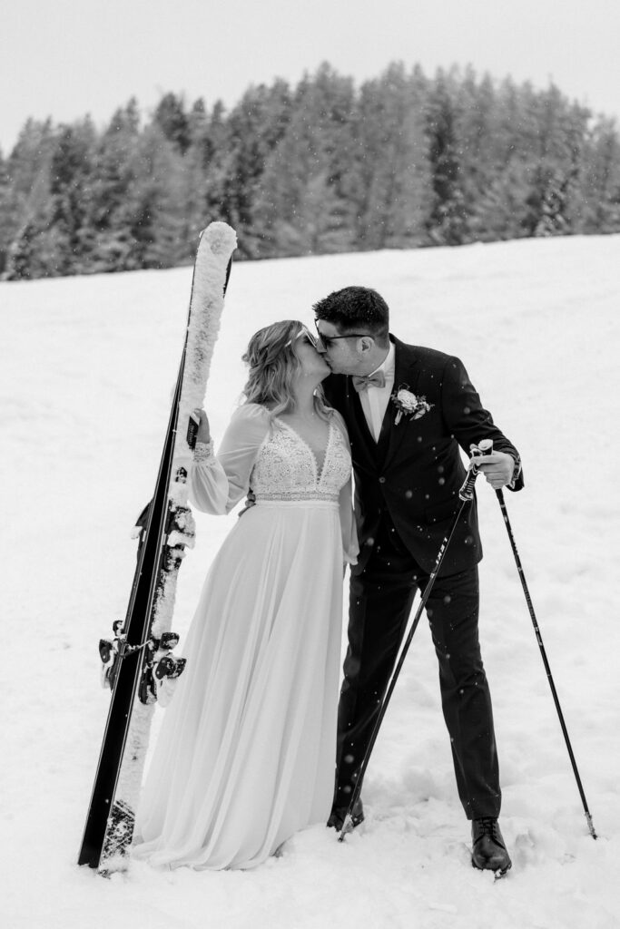 Ein Brautpaar in Hochzeitskleidung küsst sich auf verschneiten Hängen mit Skiern und Skistöcken und fängt damit den Zauber einer Berghochzeit in Tirol ein.