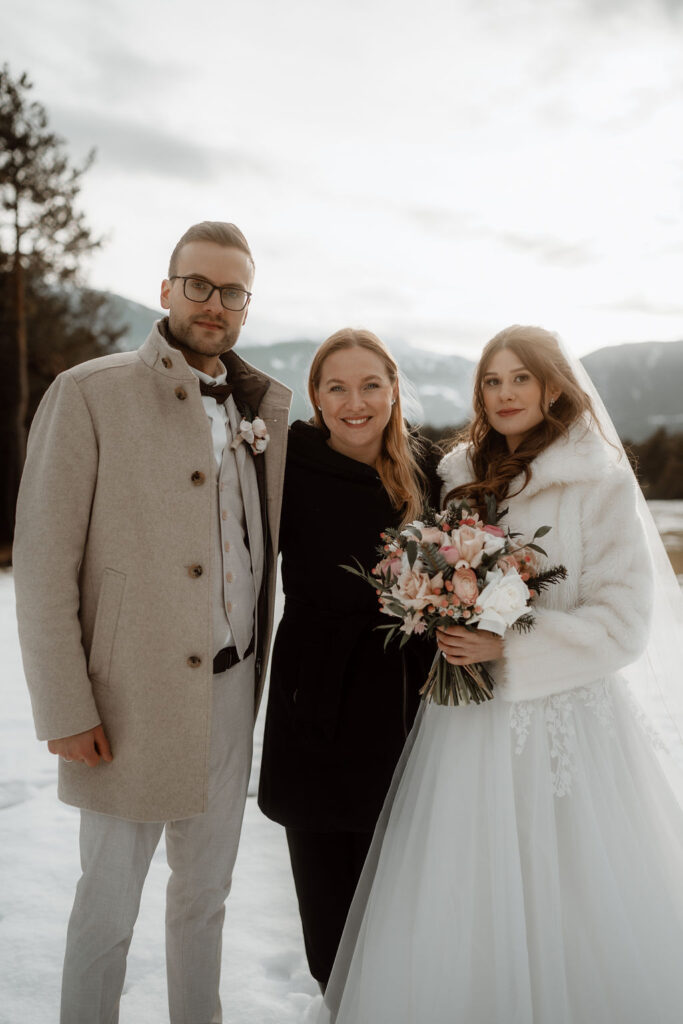 Braut und Bräutigam posieren im Freien im Schnee mit einer lächelnden Frau, Bergen und Bäumen im Hintergrund - und fangen den Zauber einer Winterhochzeit in einer atemberaubenden Berghochzeitslocation ein.