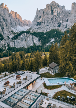 Gebirgsresort mit Sitzgelegenheiten im Freien, Pool und Pinienbäumen vor felsigen, hoch aufragenden Gipfeln.