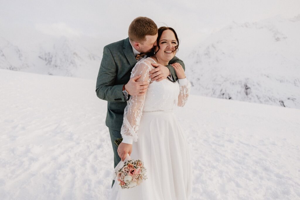 Ein Bräutigam küsst seine lächelnde Braut auf die Wange, während sie zusammen in einer verschneiten Berglandschaft stehen und ihre Winterhochzeit in der atemberaubenden Region Tirol in Österreich feiern.