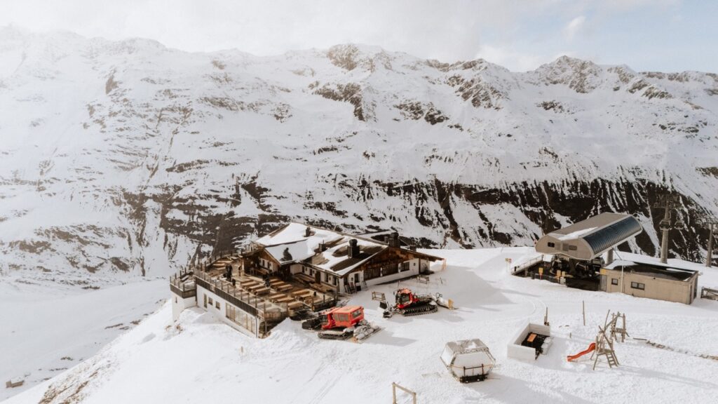 Verschneite Berghütte mit Skilift, Pistenraupen und umliegenden Gipfeln unter strahlendem Himmel - eine idyllische Hochzeitslocation in Tirol, Österreich.