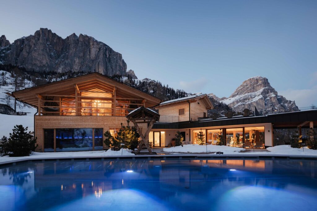 Luxuriöses Chalet mit großen Fenstern und Außenpool vor der Kulisse verschneiter Berge in der Abenddämmerung.