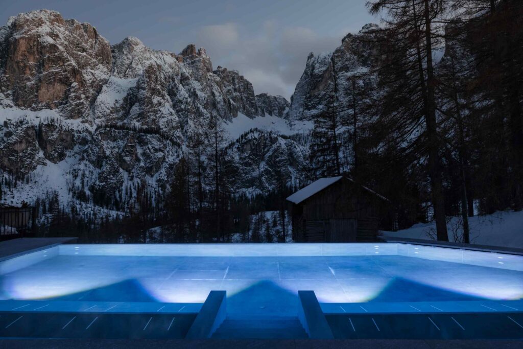 Beleuchtetes Freibad in der Abenddämmerung mit verschneiten Bergen und Kiefern im Hintergrund.