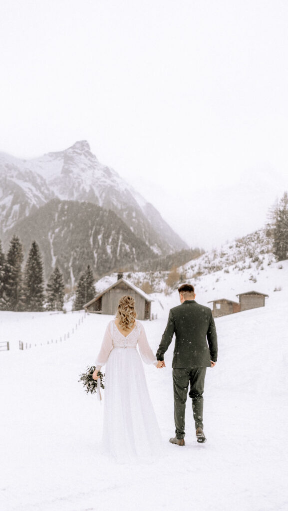 Braut und Bräutigam halten sich an den Händen und wandern in den verschneiten Bergen auf rustikale Hütten zu - die perfekte Berghochzeit für eine zauberhafte Winterhochzeit in einer verträumten Hochzeitslocation, mit dem Rücken zur Kamera.