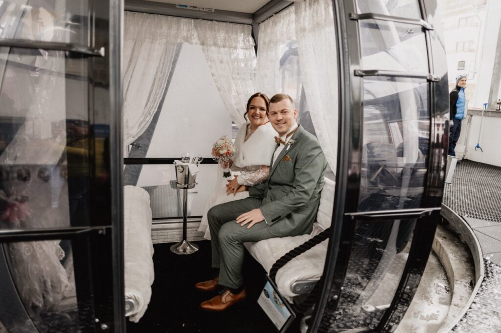 Ein lächelndes Paar in Hochzeitskleidung sitzt in einer dekorierten Riesenradkabine mit weißen Vorhängen und feiert seine Berghochzeit in einer einzigartigen Hochzeitslocation in Tirol.