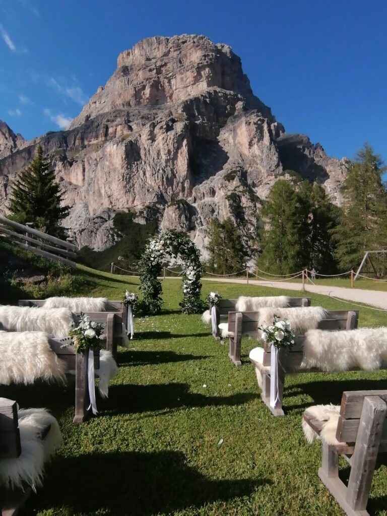 Hochzeit im Freien mit Bänken und Blumenschmuck vor einer malerischen Bergkulisse unter blauem Himmel.