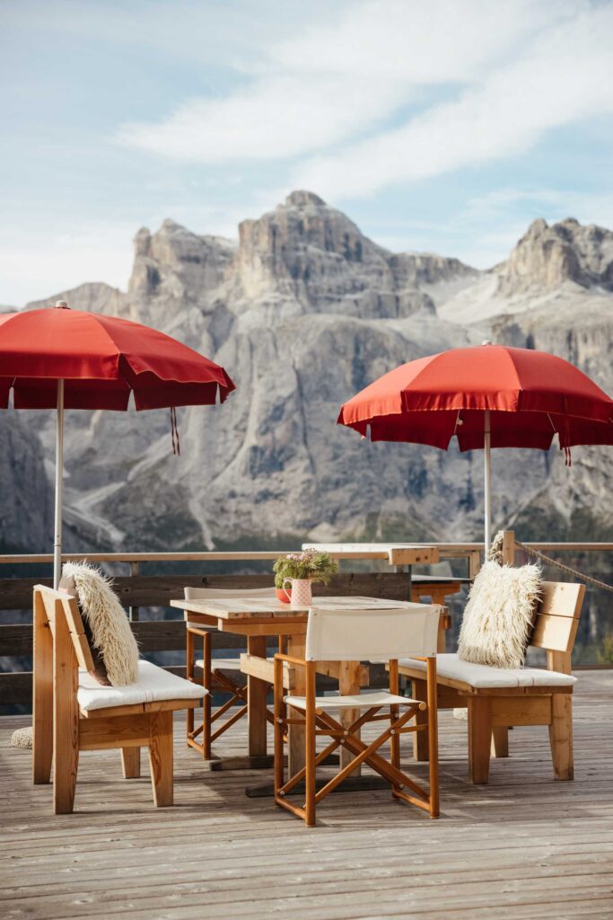 Terrassenmöbel aus Holz mit roten Sonnenschirmen auf einer Terrasse mit Blick auf felsige Berge unter blauem Himmel.