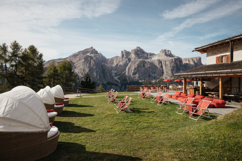 Rasenfläche mit roten Liegestühlen und weißen Schirmen, Berge im Hintergrund, neben einem rustikalen Gebäude unter blauem Himmel.