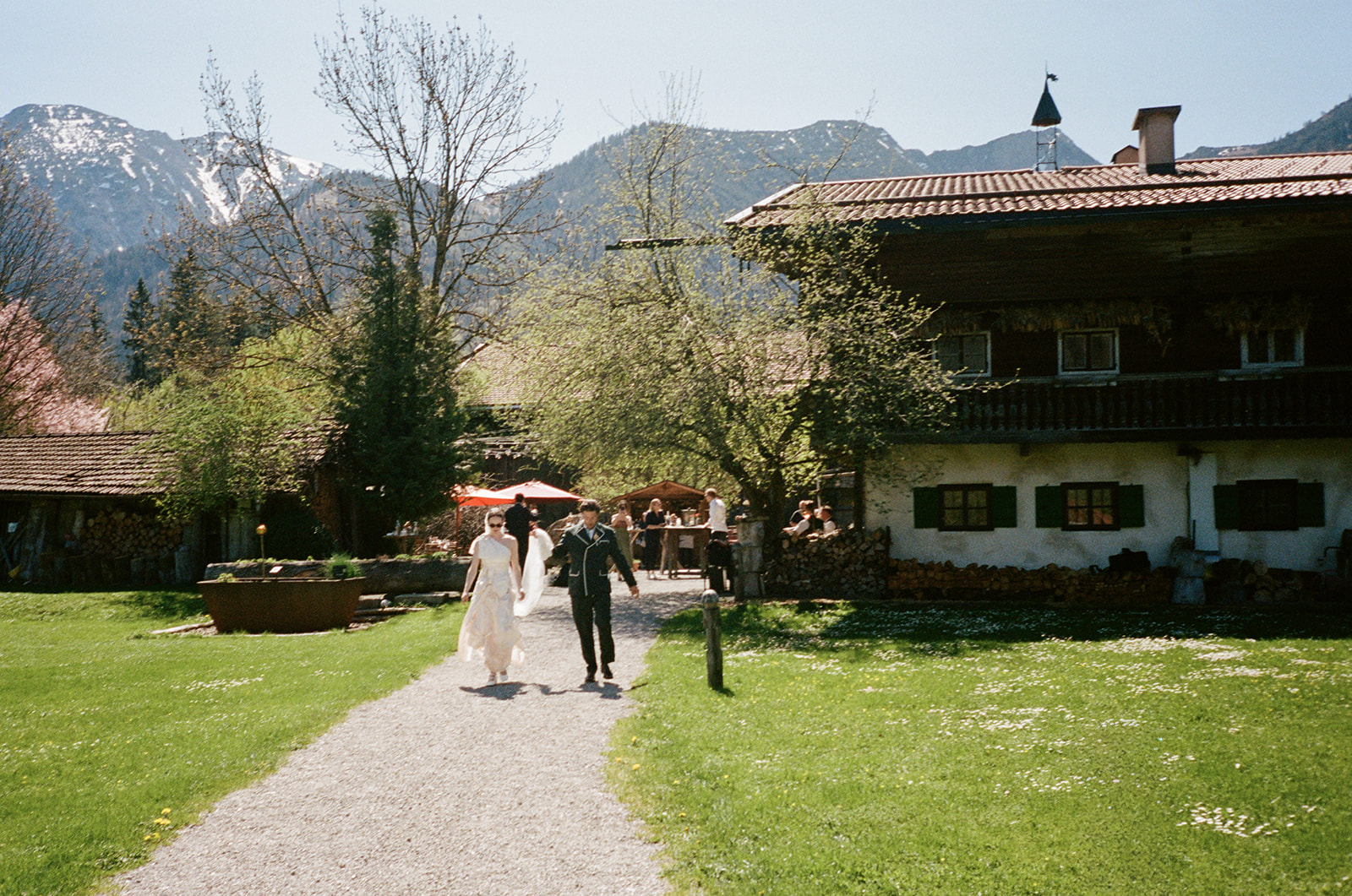 Ein Paar spaziert Hand in Hand auf einem Weg neben einem rustikalen Haus, im Hintergrund sind Berge und Bäume zu sehen – ein Bild, das den idyllischen Charme der Hochzeitslocation Hasenöhrl-Hof einfängt.