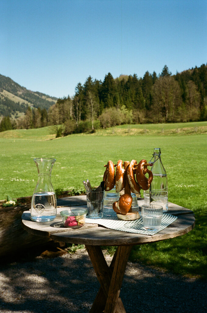 Ein runder Tisch im Freien mit Brezeln, Wasser, Gläsern und Snacks vor dem Hintergrund einer grünen Wiese und eines Waldes.