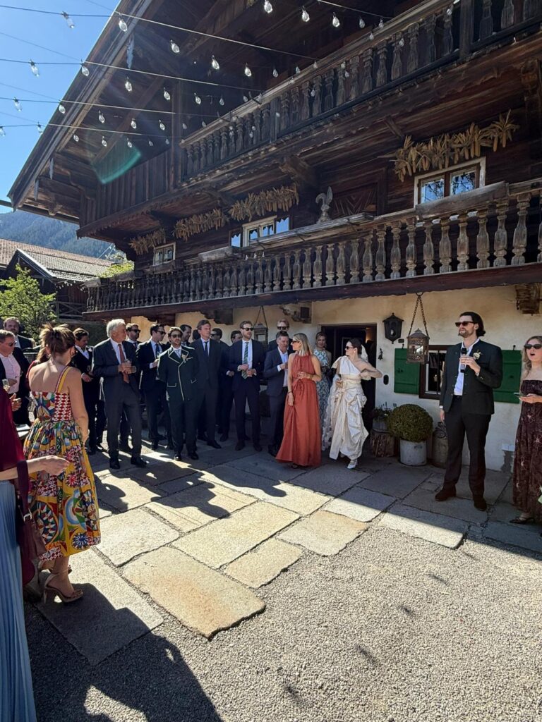 Eine Gruppe von Menschen in festlicher Kleidung steht während einer sonnigen Veranstaltung im Freien vor der rustikalen Hochzeitslocation „Hasenöhrl-Hof“ aus Holz.