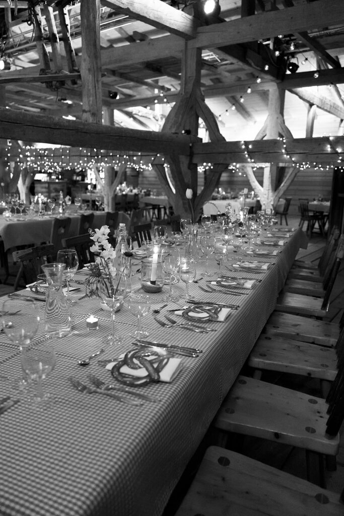 Langer Banketttisch mit karierter Tischdecke, gedeckt für ein Abendessen in einer rustikalen Scheune mit funkelnden Lichterketten.