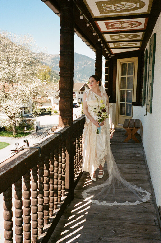 Eine Braut in einem cremefarbenen Kleid steht auf einem Holzbalkon, hält einen Blumenstrauß in der Hand und im Hintergrund sind Berge zu sehen.